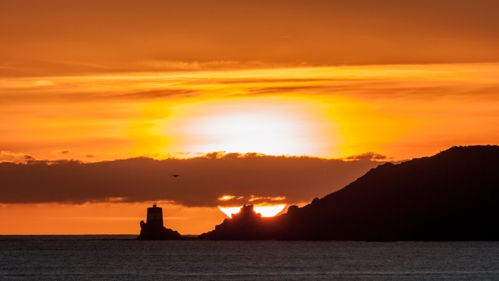 Seascape showing a big orange sunset on a long zoom behind Noirmont Tower, taken from the promenade in front of West Park, St. Helier, Jersey, Channel Islands