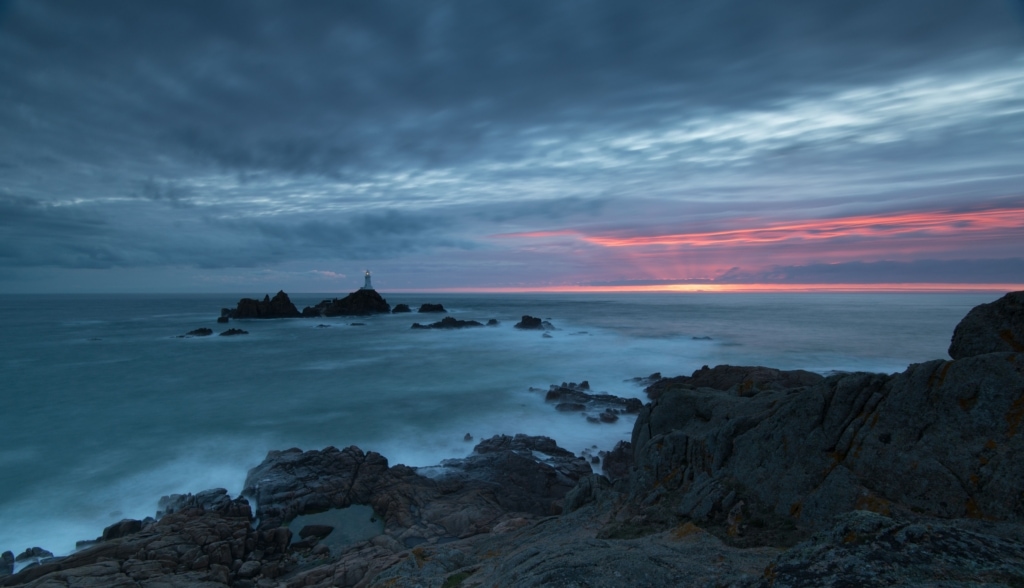 Long exposure of the sea and clouds and rocks and Corbiere Lighthouse at sunset and at high tide, taken from above and behind the bottom car park at Corbiere, St. Brelade, Jersey, Channel Islands