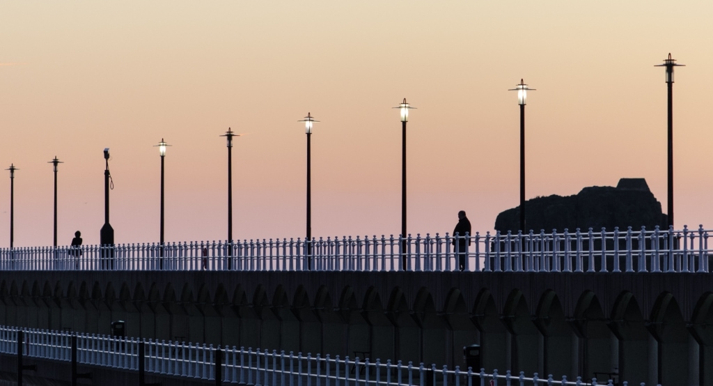 Fat man looking at fit girl taking a walk at St. Helier Marina at sunset with St. Helier Hermitage in the background, St. Helier, Jersey, Channel Islands