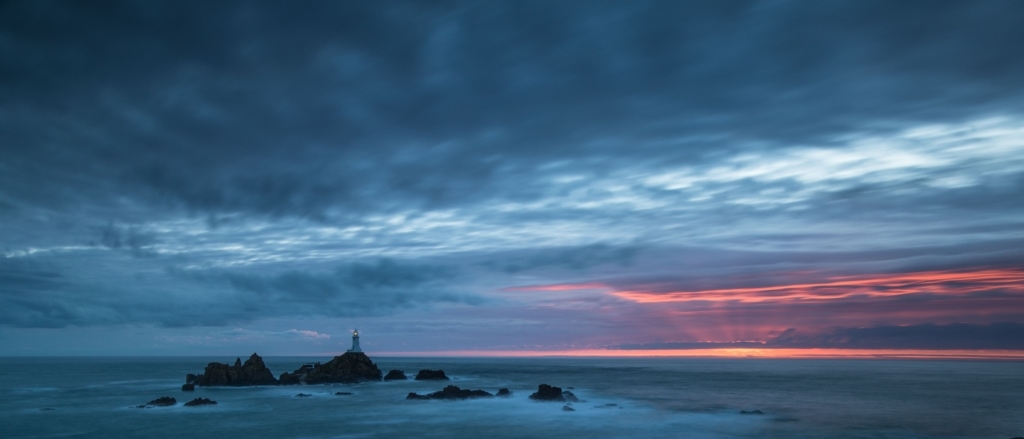 Long exposure of the sea and clouds and rocks and Corbiere Lighthouse at sunset and at high tide, taken from above and behind the bottom car park at Corbiere, St. Brelade, Jersey, Channel Islands