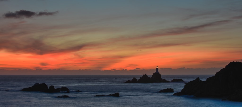 Silhouette of Corbiere Lighthouse and cliffs with vibrant clouds at sunset from La Rosiere, St. Brelade, Jersey, Channel Islands