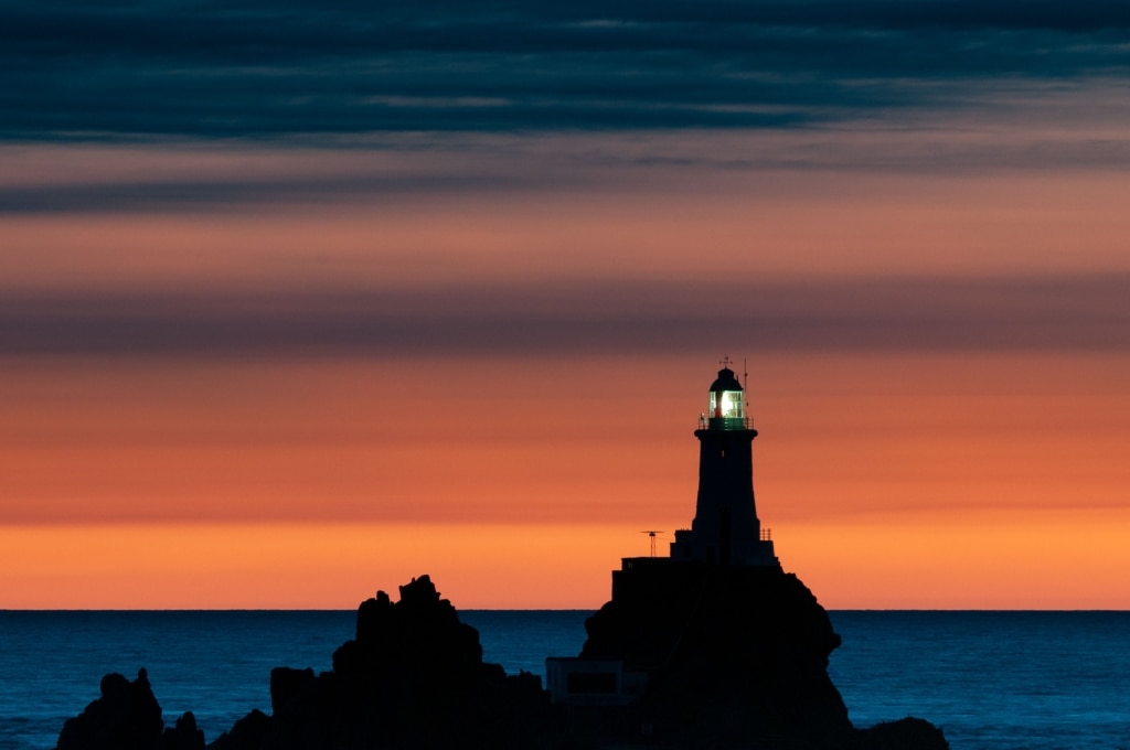 Silhouette of illuminated Corbiere Lighthouse with vibrant clouds at sunset, from La Rosiere, St. Brelade, Jersey, Channel Islands