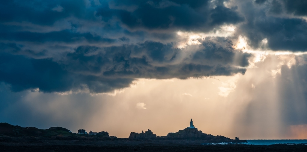 Sun rays and stormy clouds above Corbiere Lighthouse from Le Braye Slip, St. Brelade, Jersey, Channel Islands