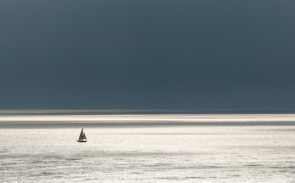 Tiny sailing boat far out to sea with heavy rain clouds moving away beyond the horizon, taken at Corbiere Phare, St. Brelade, Jersey, Channel Islands