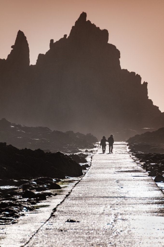 Walking couple and silhouetted rocks at the end of Corbiere Lighthouse causeway, St. Brelade, Jersey, Channel Islands